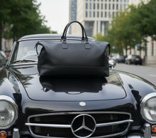 Load image into Gallery viewer, A Franklin-Christoph Duffle Bag - Black Leather sits on a classic black Mercedes-Benz, ready for a stylish getaway.