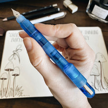 Load image into Gallery viewer, Hand holding a Franklin-Christoph Model 25 Eclipse Fountain Pen in Maya Blue above a notebook with mushroom drawings.