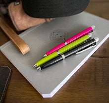 Load image into Gallery viewer, Three pens, including a Franklin-Christoph Model 28 Shortstock Ballpoint, rest on a gray notebook near a bag and hat.