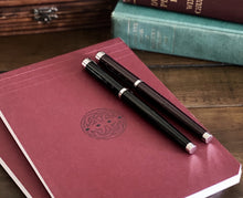 Load image into Gallery viewer, Two Franklin-Christoph Model 28 Libertas fountain pens rest on a burgundy Celtic-emblem notebook beside books.