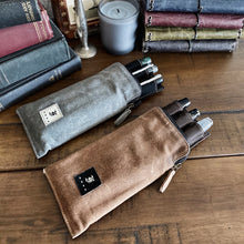 Load image into Gallery viewer, Two Franklin-Christoph pen cases with Pen Case Inserts rest near stacked notebooks and a gray candle on a wood table.