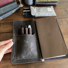 Load image into Gallery viewer, A hand holds a Franklin-Christoph Pen Case Insert in a leather notebook cover on a wooden table with books and journal.