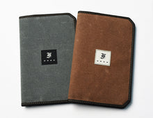 Load image into Gallery viewer, Two rectangular, fabric-covered pocket notebooks are displayed against a white background. One is grey with a black label, and the other is brown with a white label. Both feature a black stitched border and a central patch displaying an ornate F logo. These refined notebooks are finished with fountain pen friendly paper and evoke the timeless durability of a waxed canvas notebook cover.