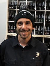 Load image into Gallery viewer, A man wearing a Franklin-Christoph F-C Beanie smiles in front of shelves stocked with tattoo ink bottles.