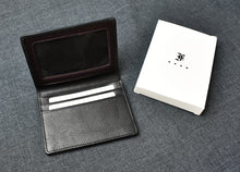 Load image into Gallery viewer, A Franklin-Christoph Card Wallet crafted from black grained napa leather, featuring multiple card slots and a clear ID card slot, is displayed open beside a white box bearing a black symbol on a dark grey fabric surface.