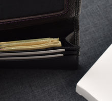 Load image into Gallery viewer, An open Franklin-Christoph Card Wallet, crafted from grained napa leather in black, displays folded cash and two cards inside—one placed in the dedicated ID slot—as it rests on a dark fabric surface beside a white rectangular object.