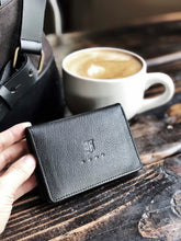 Load image into Gallery viewer, A hand holds a Franklin-Christoph Card Wallet crafted from black grained napa leather near a cup of coffee on a rustic wooden table, with part of a gray bag visible in the background. The wallet features a discreet ID card slot for added convenience.