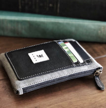 Load image into Gallery viewer, The Franklin-Christoph Zippered Card Wallet, made from supple napa leather, features a black front pocket with a stitched white logo and multiple card slots. It rests on a wooden surface, with blurred books visible in the background.