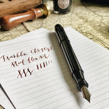 Load image into Gallery viewer, Franklin-Christoph #6 Nib Unit gold fountain pen on lined paper with calligraphy and ink bottle in the background.