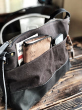 Load image into Gallery viewer, Franklin-Christoph Fortis Sling Bag in Iron Brown and Black sits on a rustic table with a book and notebook in front pocket.