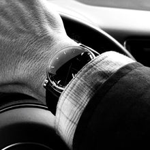 Load image into Gallery viewer, A hand wearing the Franklin-Christoph IWO Timepiece grips a car’s wheel in a black-and-white photo.