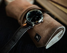 Load image into Gallery viewer, A Franklin-Christoph IWO Timepiece with a black leather strap sits on a brown suede roll next to a closed book.