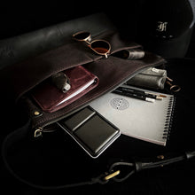 Load image into Gallery viewer, A Franklin-Christoph Fortis Sling Bag in Iron Brown/Black holds a maroon notebook, sketchbook, and art supplies.