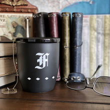 Load image into Gallery viewer, The Franklin-Christoph Coffee Mug 1 is matte black with a white gothic F and diamond accents, shown on a desk with books.