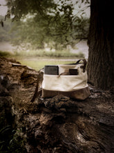 Load image into Gallery viewer, A Franklin-Christoph Fortis Sling Bag - Creme’ and Brown, packed with books and a wallet, rests on a fallen tree.