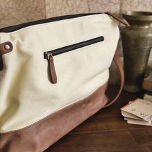 Load image into Gallery viewer, A Franklin-Christoph Fortis Sling Bag - Creme’ and Brown rests by a metal vase and a stack of playing cards.