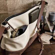 Load image into Gallery viewer, A Franklin-Christoph Fortis Sling Bag - Creme’ and Brown rests on a desk with playing cards and notebooks nearby.