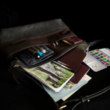 Load image into Gallery viewer, Open the Franklin-Christoph Fortis Sling Bag - Iron Brown and Black, filled with notebooks, pens, brushes, paints, and art.