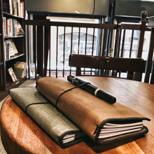 Load image into Gallery viewer, Two journals and a fountain pen rest on a table—one is a Franklin-Christoph Vagabond Notebook Cover—books behind.