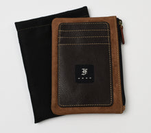 Load image into Gallery viewer, The Franklin-Christoph Zippered Card Wallet showcases a two-toned napa leather design, with a tan suede border framing a dark brown pebbled center equipped with card slots and a side zipper. A small black square patch featuring the signature gothic-style F logo embellishes the front, and the wallet is presented on a black fabric pouch.