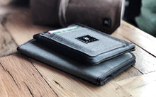 Load image into Gallery viewer, A Franklin-Christoph Zippered Card Wallet in grey fabric, with partially visible cards in its slots, rests atop a matching notebook on a wooden table.