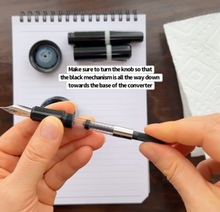 Load image into Gallery viewer, Hands holding a Franklin-Christoph Fountain Pen Converter and pen above a notebook, cap and pen on the table.