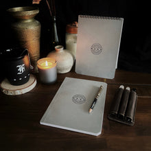 Load image into Gallery viewer, A candle, Franklin-Christoph Greypads spiral pad, pen, and pottery sit on a desk; leather pen case is nearby.
