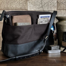 Load image into Gallery viewer, A Franklin-Christoph Fortis Sling Bag - Iron Brown and Black rests on a desk with maps, wallets, and a vintage vase.