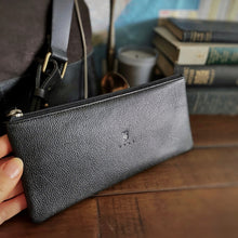 Load image into Gallery viewer, A hand holds the Franklin-Christoph Currency Case - Leather on a wooden table with books and a bag in the background.
