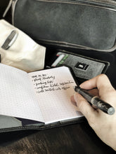Load image into Gallery viewer, A person’s hand uses a black fountain pen to write in a dotted journal with fountain pen friendly paper. In the background, there is a travel bag, wallet, and a Franklin-Christoph 5.3 Pocket Notebook Cover - Canvas.