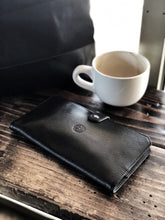 Load image into Gallery viewer, A Franklin-Christoph ETN - Black Napa w/ Suit Grey rests beside a white coffee cup on a rustic window-side table.