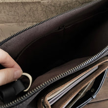 Load image into Gallery viewer, Hand unzips a Franklin-Christoph Fortis Sling Bag in Iron Brown/Black, showing organized pockets and papers inside.