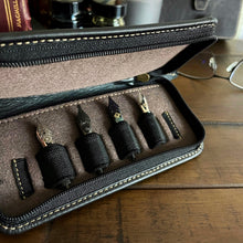 Load image into Gallery viewer, A Franklin-Christoph 4 Nib Case holds four nibs with black grips on a wooden table beside eyeglasses.
