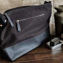 Load image into Gallery viewer, An Iron Brown and Black Fortis Sling Bag by Franklin-Christoph rests on wood near a vase and metal tin.