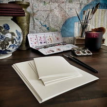 Load image into Gallery viewer, A desk with Franklin-Christoph Water Color Paper - Loose Leaf, brushes, a candle, a map, and a decorative vase.