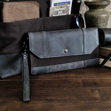 Load image into Gallery viewer, A Franklin-Christoph Fortis Clutch Envelope - Iron Brown &amp; Black with a wrist strap rests on a table with travel items.